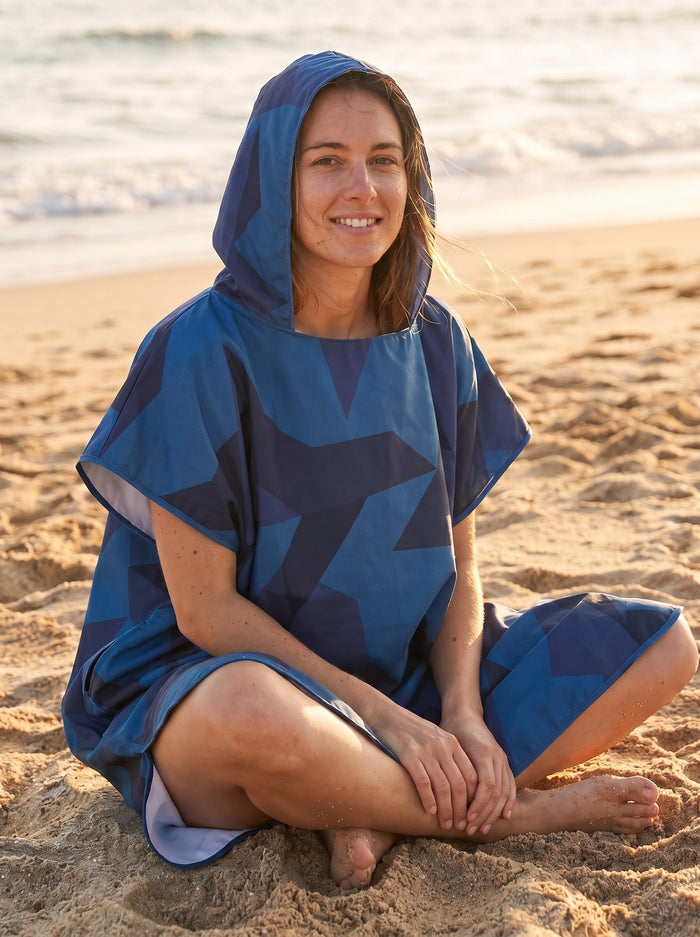 Femme assise sur le sable en poncho serviette à capuche bleu, face à la mer.