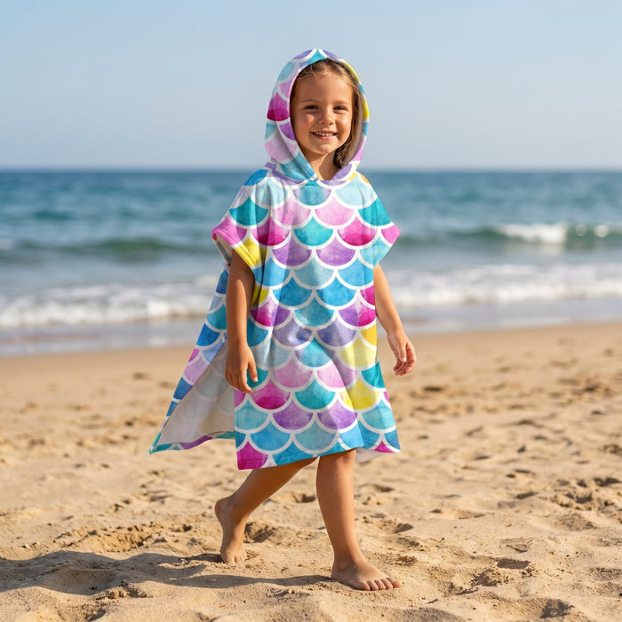 Poncho serviette à capuche motif écailles multicolores, porté par une enfant souriante marchant sur le sable.