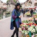 Poncho cape colorblock vert et violet porté par une femme avec un chapeau, regardant des fleurs au marché.