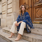 Poncho cape femme à motif géométrique et franges bleu et beige, porté par une femme assise sur des marches en pierre