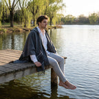 Poncho cape homme à carreaux avec poches gris, porté par un homme assis de profil sur un ponton au bord de l'eau