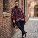 Poncho cape homme paisley bordeaux, porté par un homme de profil, appuyé contre un mur de briques dans une ruelle
