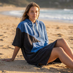 Poncho serviette à capuche bicolore bleu et noir, porté par une femme assise sur le sable.
