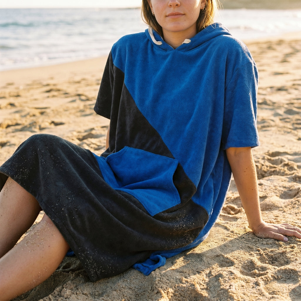 Poncho serviette à capuche bicolore bleu et noir, porté par une femme assise sur le sable, vue de trois quarts.