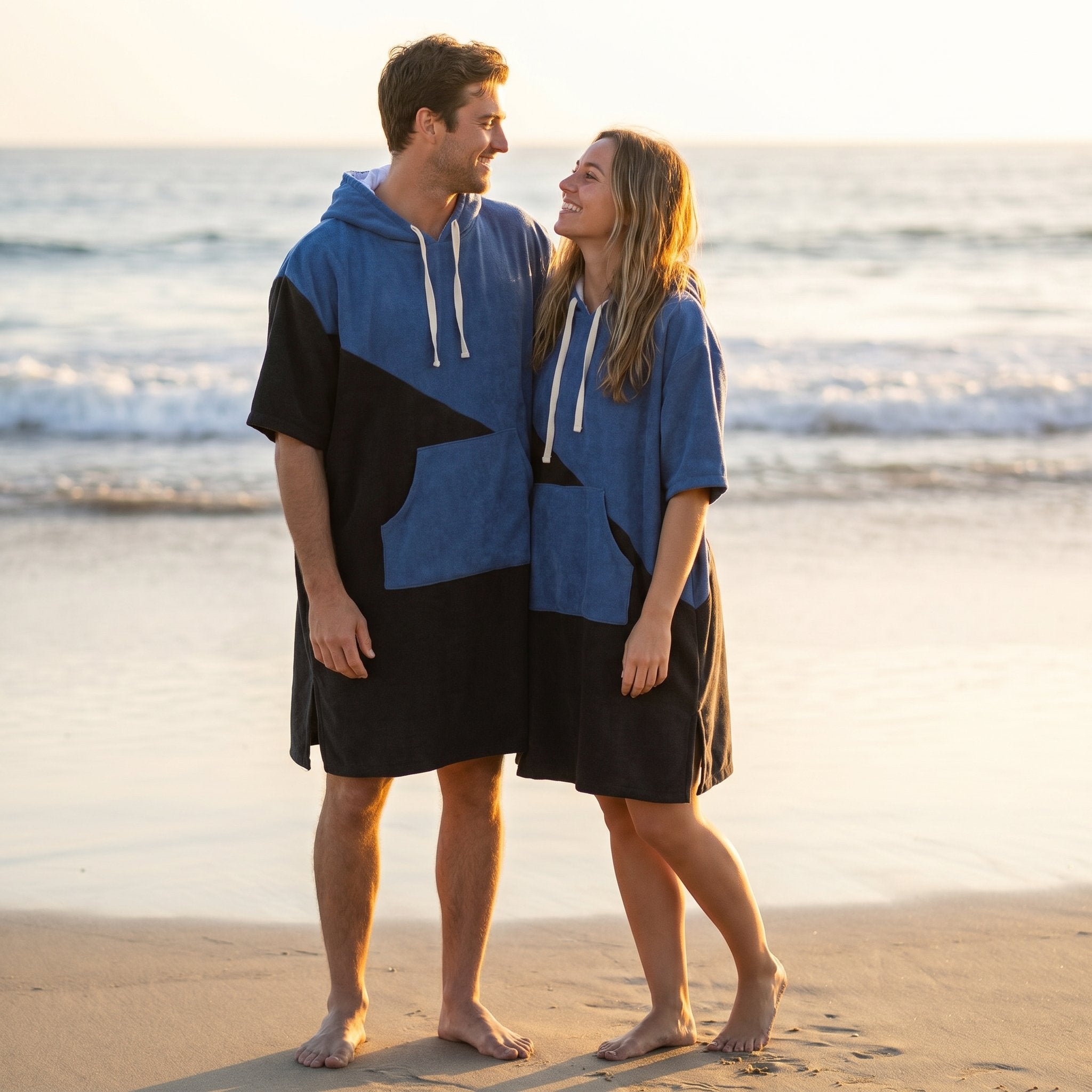 Poncho serviette à capuche bicolore bleu et noir, porté par un homme et une femme debout sur la plage.