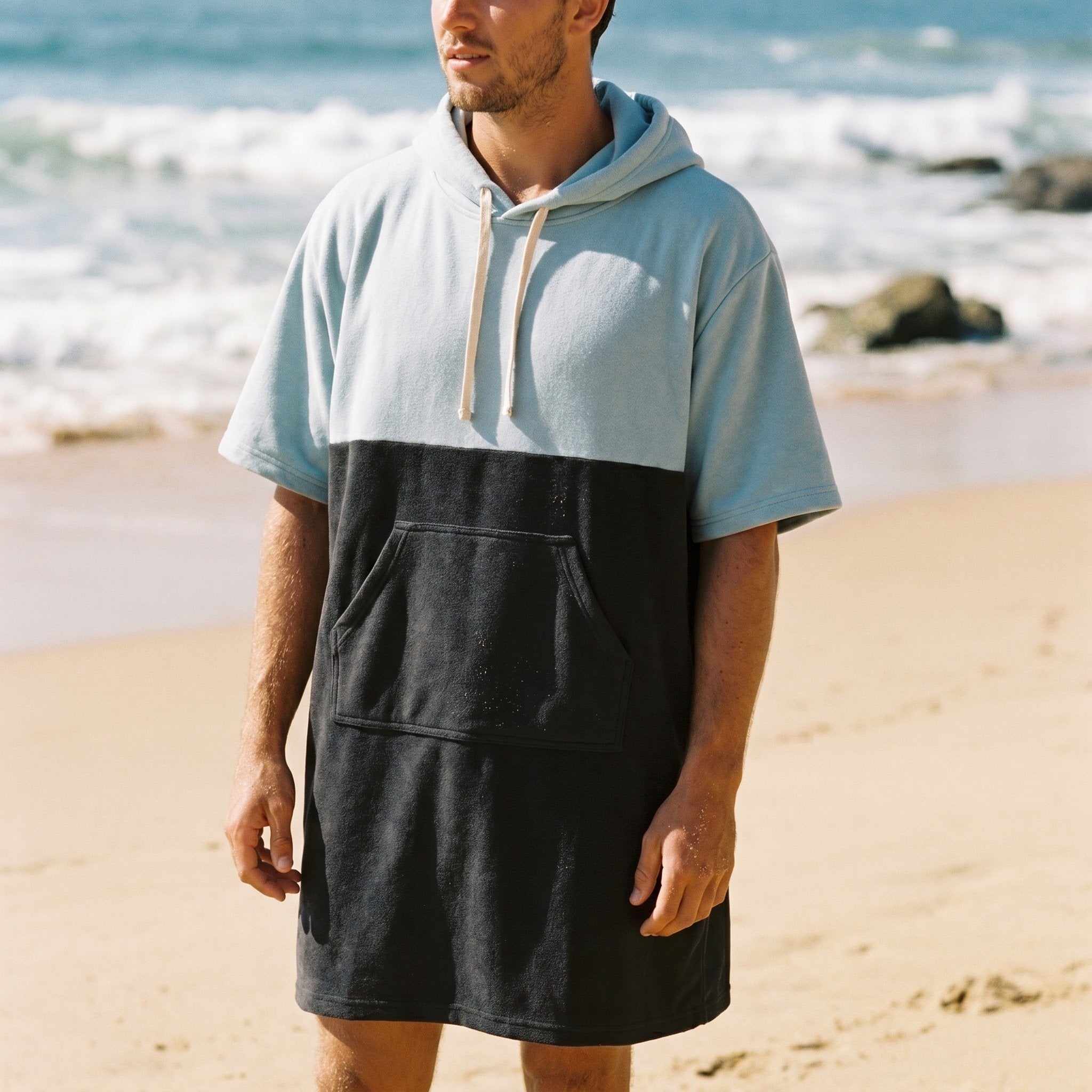 Poncho serviette à capuche bicolore ciel et gris, porté par un homme debout sur la plage, vue de face en plan américain.