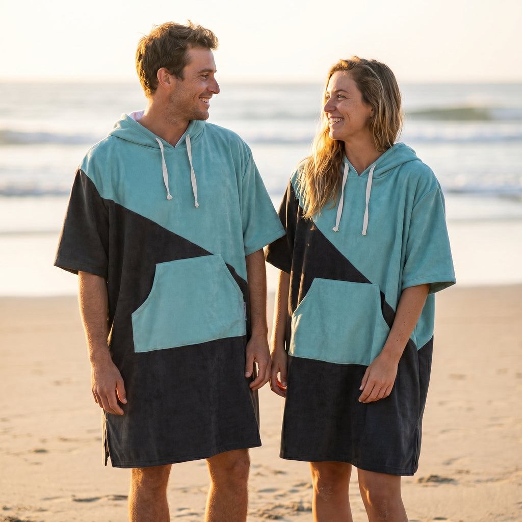 Poncho serviette à capuche bicolore turquoise et gris porté par un homme et une femme se faisant face sur la plage.