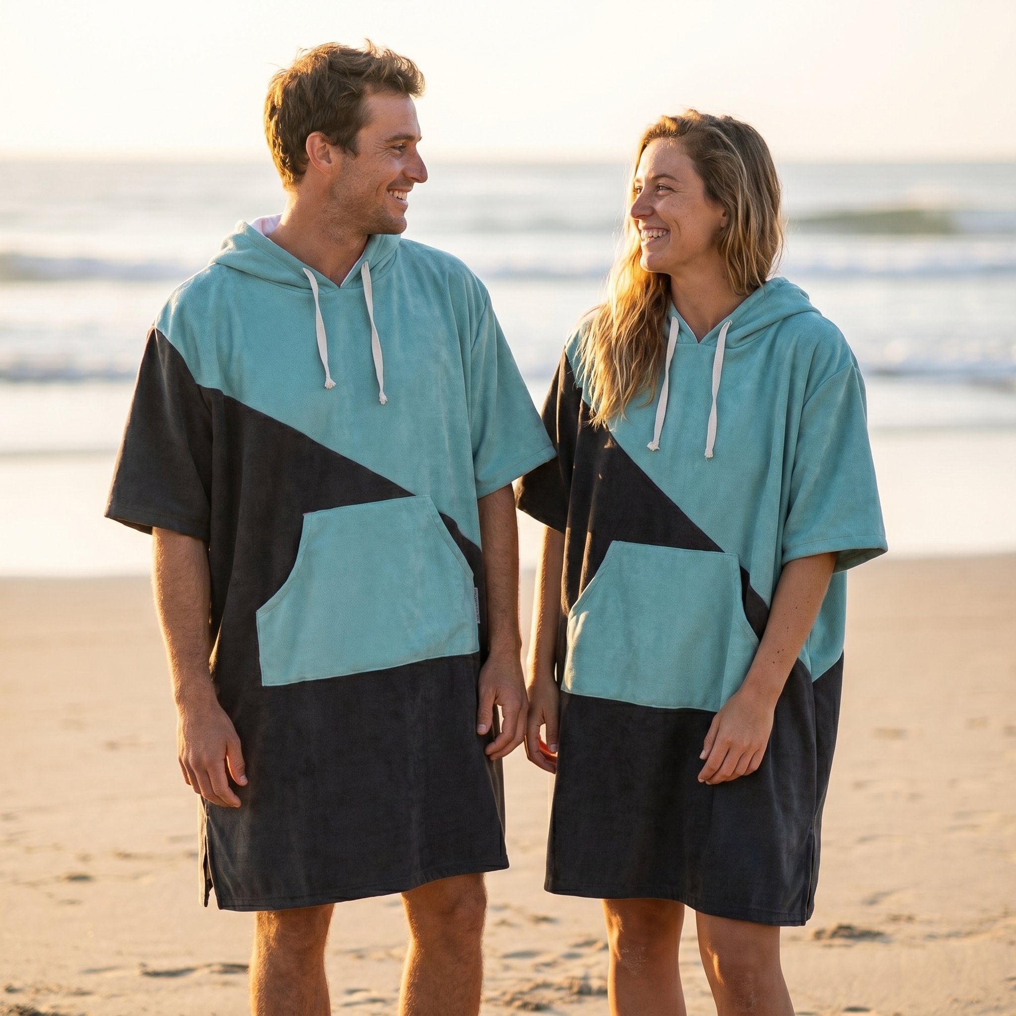 Poncho serviette à capuche bicolore turquoise et gris porté par un homme et une femme se faisant face sur la plage.