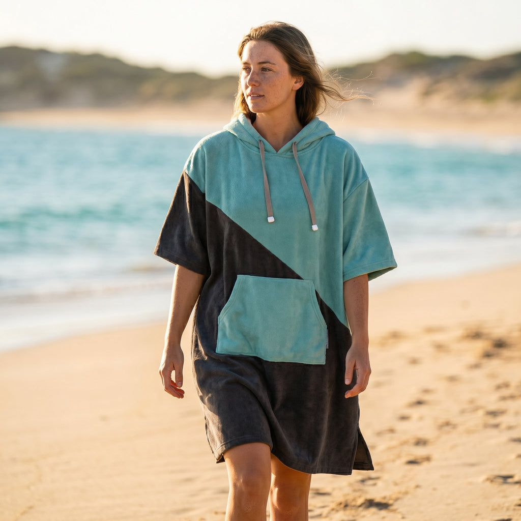 Poncho serviette à capuche bicolore turquoise et gris porté par une femme marchant de profil sur le sable au bord de l'eau.