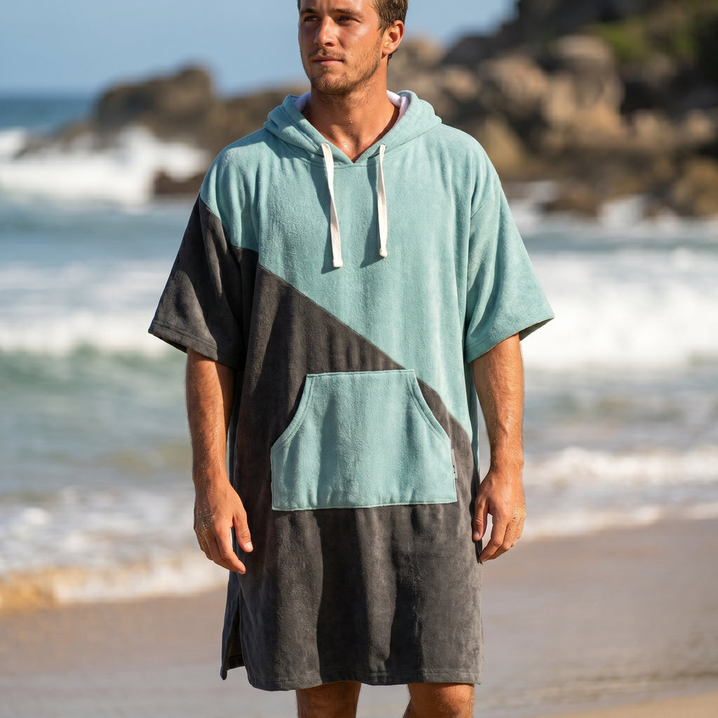 Poncho serviette à capuche bicolore turquoise et gris porté par un homme debout de face sur une plage rocheuse.