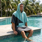 Poncho serviette à capuche bicolore turquoise et gris porté par un homme assis au bord d'une piscine, les pieds dans l'eau.