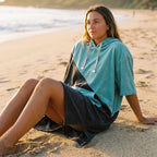 Poncho serviette à capuche bicolore turquoise et gris porté par une femme assise sur le sable, regardant vers l'horizon.