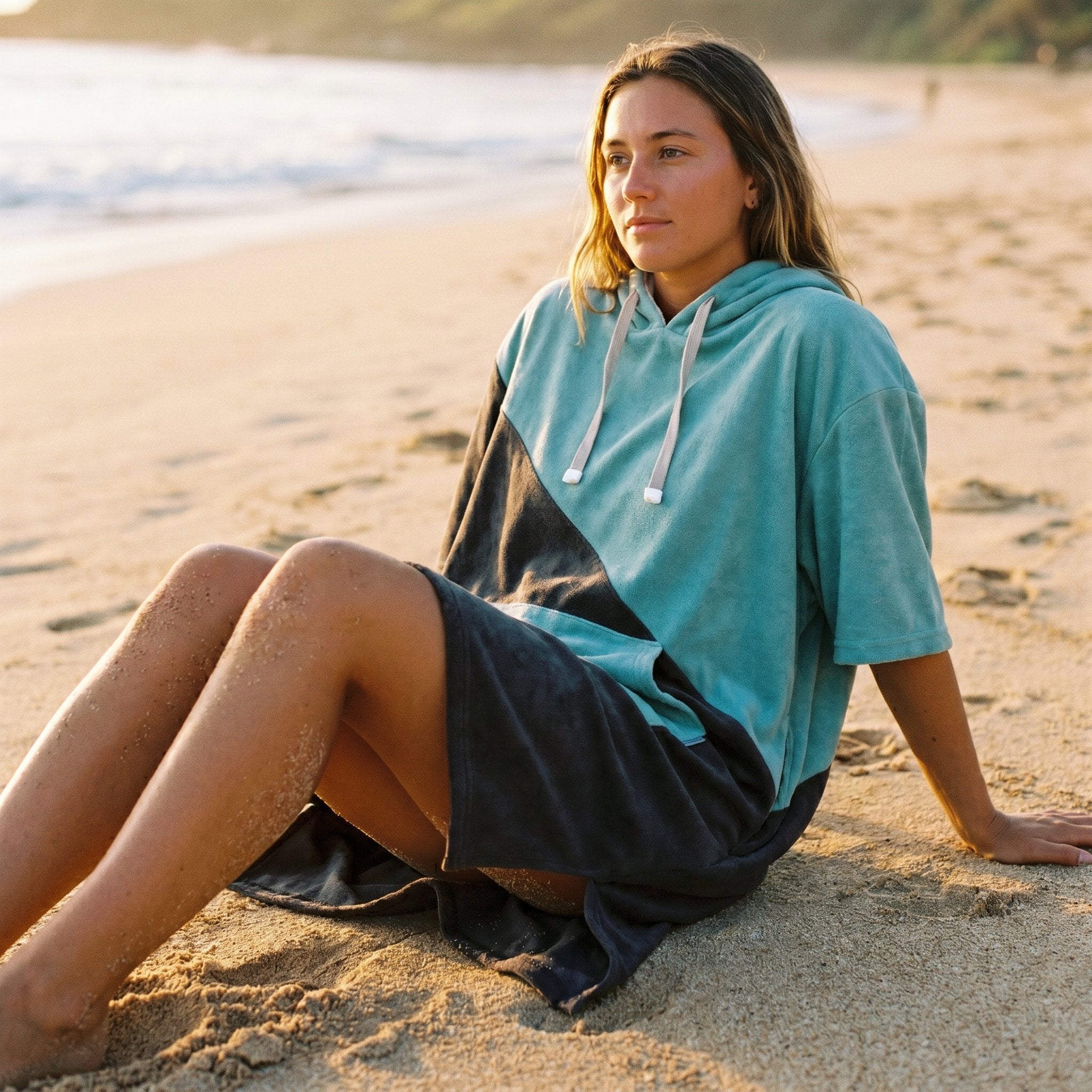 Poncho serviette à capuche bicolore turquoise et gris porté par une femme assise sur le sable, regardant vers l'horizon.