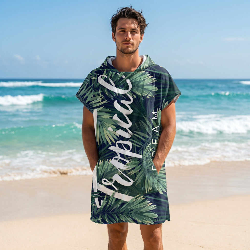 Poncho serviette à capuche motif feuillage tropical, porté par un homme sur une plage de sable fin.