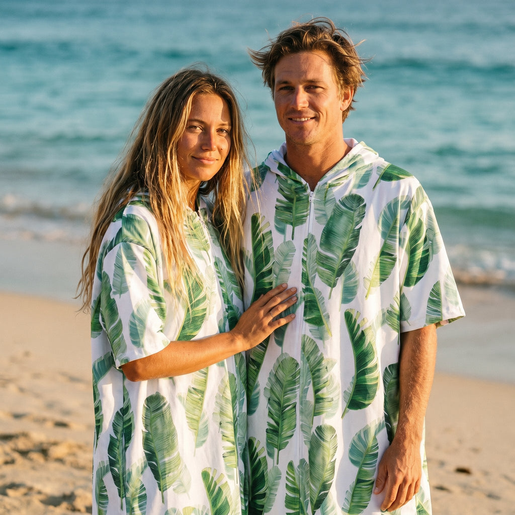 Poncho serviette a capuche motif feuilles de bananier porté par un couple souriant sur une plage ensoleillée.