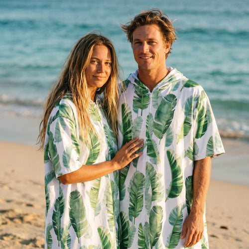Poncho serviette a capuche motif feuilles de bananier porté par un couple souriant sur une plage ensoleillée.