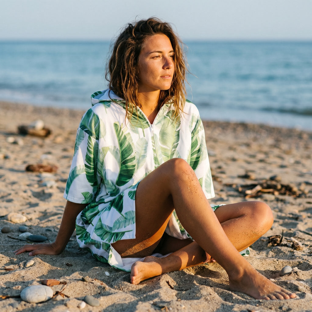 Poncho serviette a capuche motif feuilles de bananier porté par une femme assise sur le sable, regardant vers l'horizon.