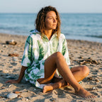 Poncho serviette a capuche motif feuilles de bananier porté par une femme assise sur le sable, regardant vers l'horizon.