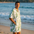 Poncho serviette a capuche motif feuilles de bananier porté par un homme souriant, debout sur la plage face à la mer.