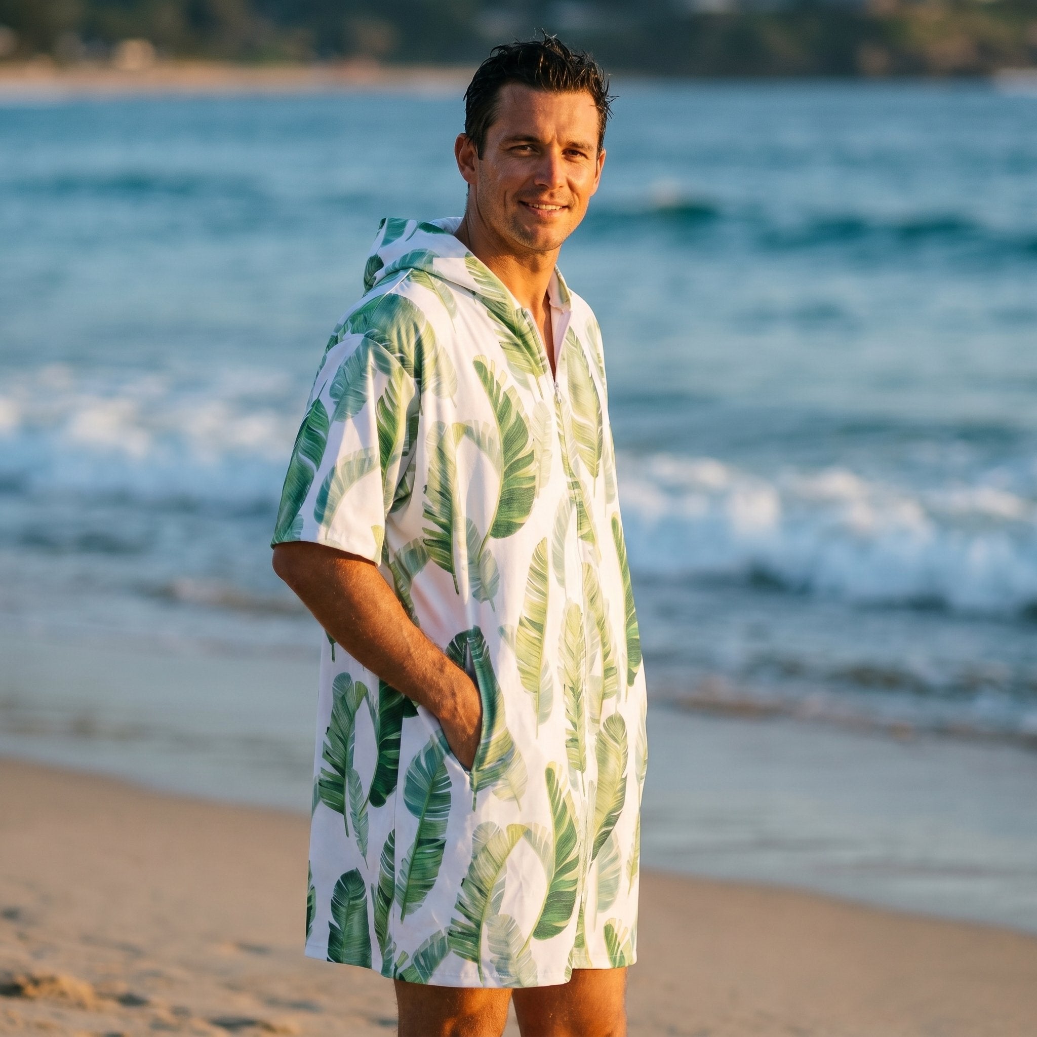 Poncho serviette a capuche motif feuilles de bananier porté par un homme souriant, debout sur la plage face à la mer.
