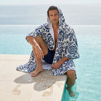 Poncho serviette à capuche motif fleurs bleues, porté par un homme assis au bord d'une piscine.