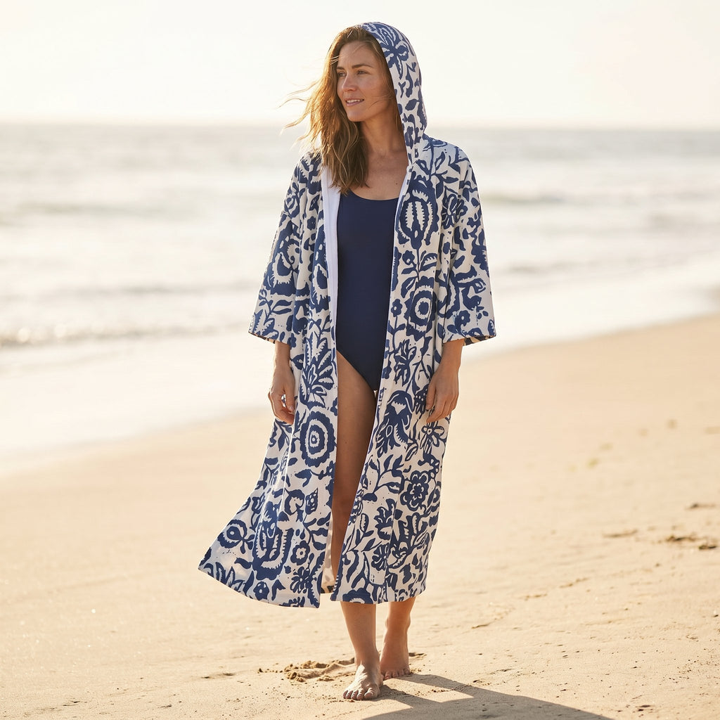 Poncho serviette à capuche motif fleurs bleues, porté par une femme marchant sur le sable.