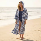 Poncho serviette à capuche motif fleurs bleues, porté par une femme marchant sur le sable.