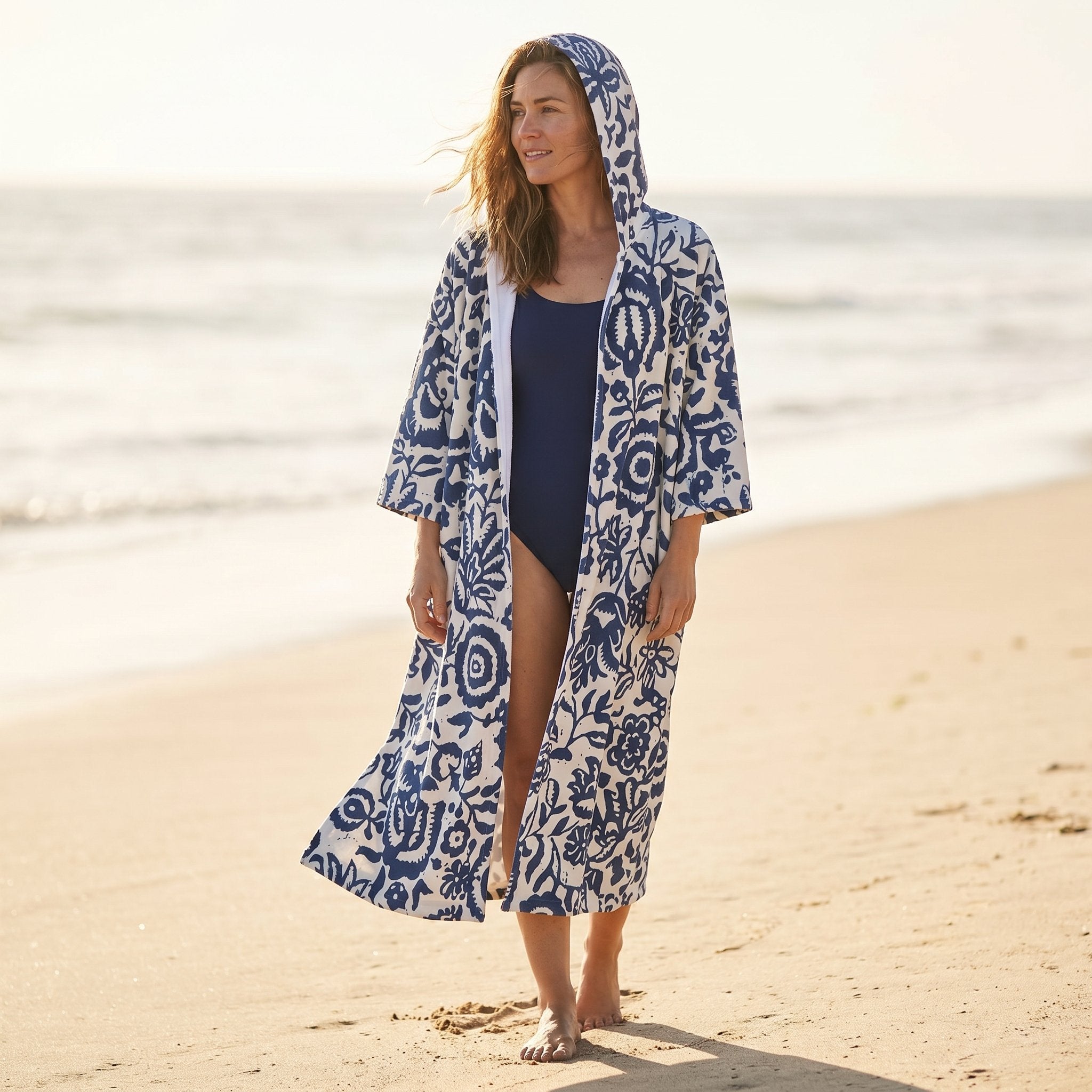 Poncho serviette à capuche motif fleurs bleues, porté par une femme marchant sur le sable.