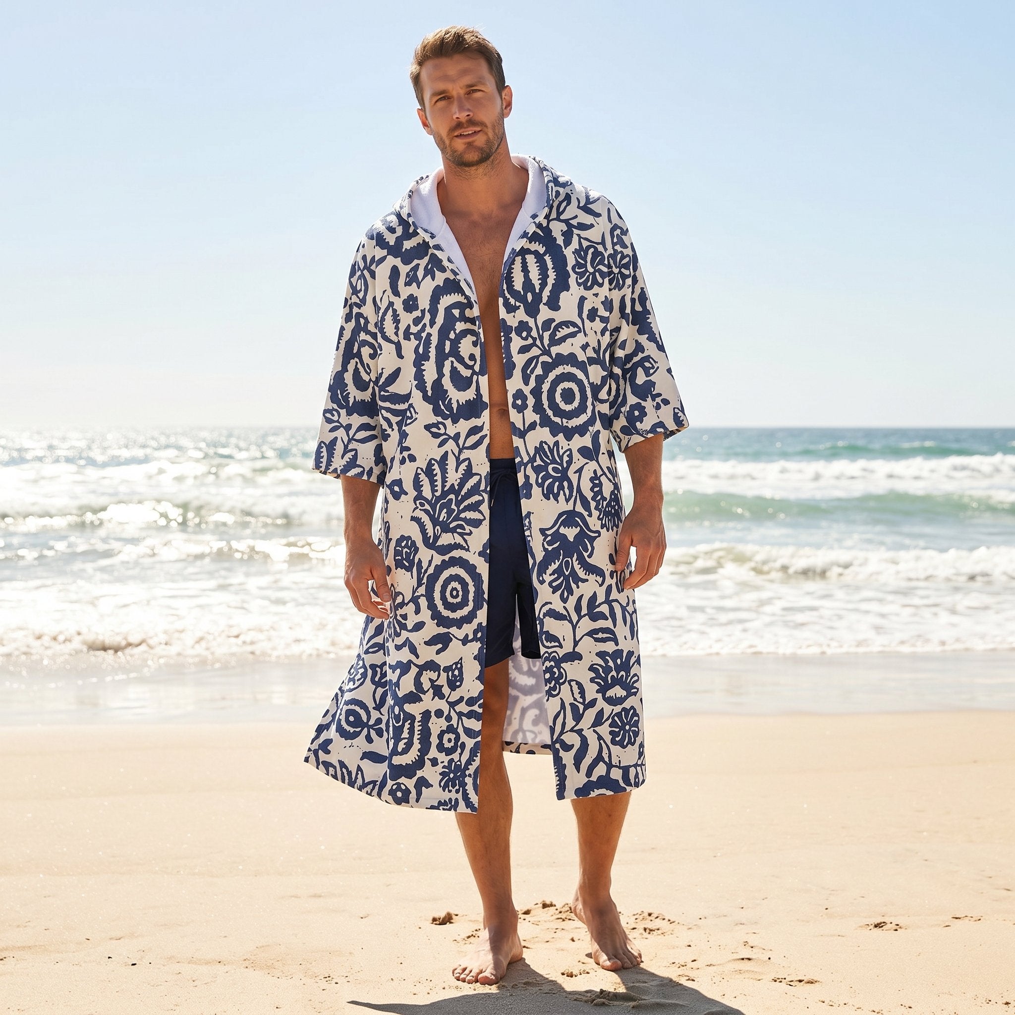 Poncho serviette à capuche motif fleurs bleues, porté par un homme debout sur la plage.