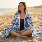 Poncho serviette à capuche motif fleurs bleues, porté par une femme assise en tailleur sur le sable.
