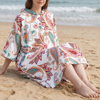Poncho serviette a capuche motif paisley colore, porte par une femme assise sur le sable.