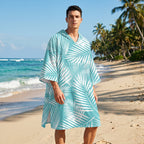 Poncho serviette à capuche motif palmes turquoise, porté par un homme debout sur une plage de sable fin.