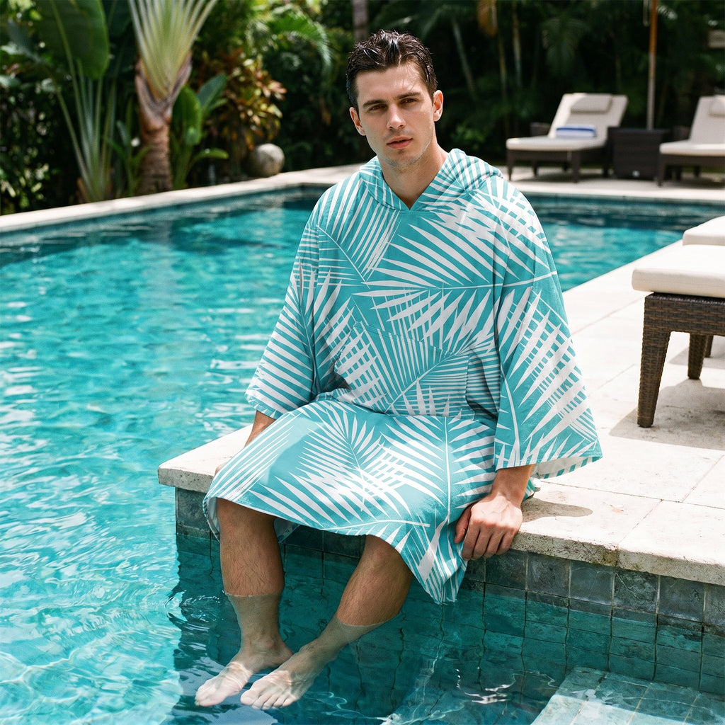 Poncho serviette à capuche motif palmes turquoise, porté par un homme assis au bord d'une piscine, les pieds dans l'eau.