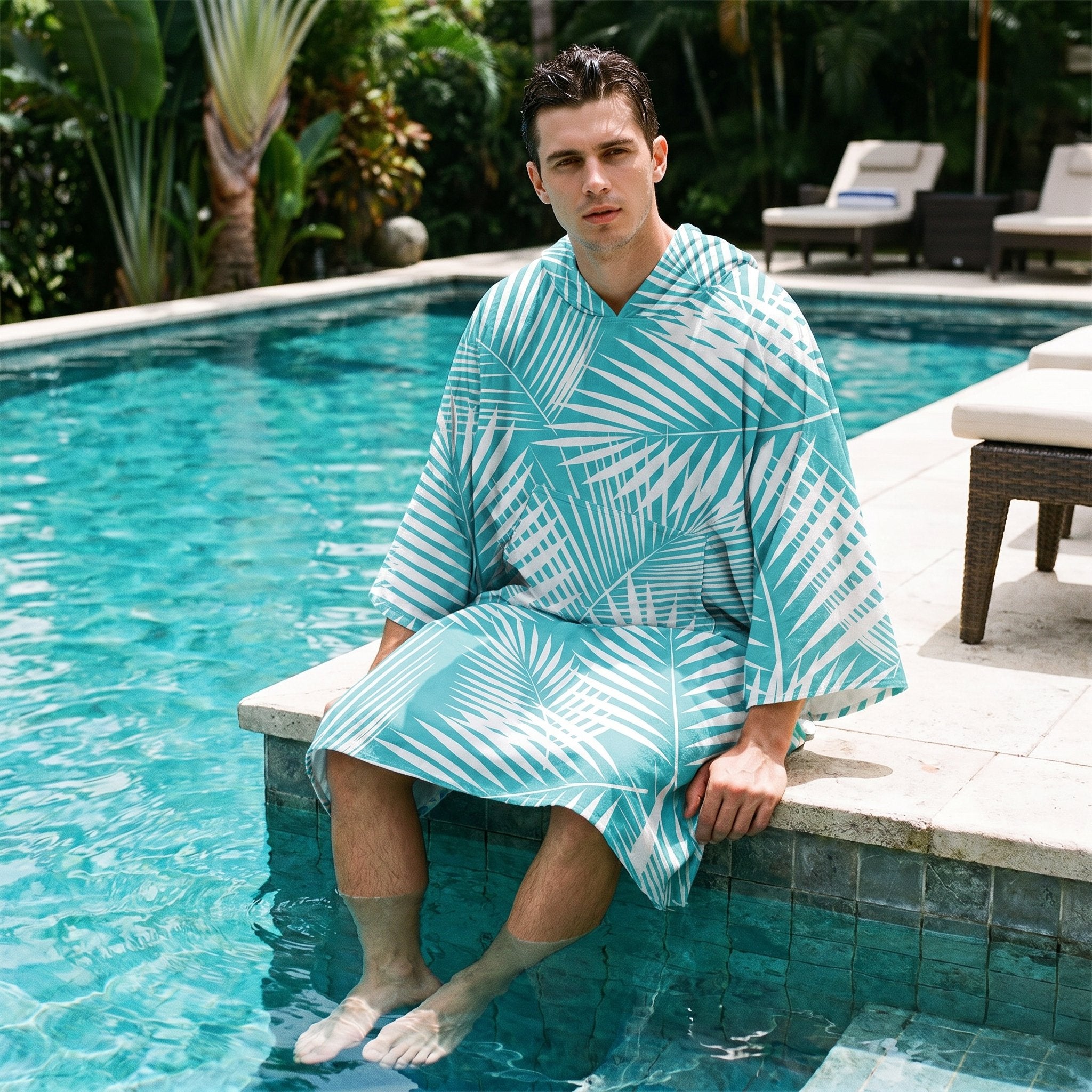 Poncho serviette à capuche motif palmes turquoise, porté par un homme assis au bord d'une piscine, les pieds dans l'eau.