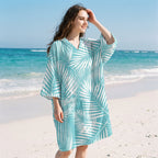 Poncho serviette à capuche motif palmes turquoise, porté par une femme souriante, debout sur le sable face à la mer.