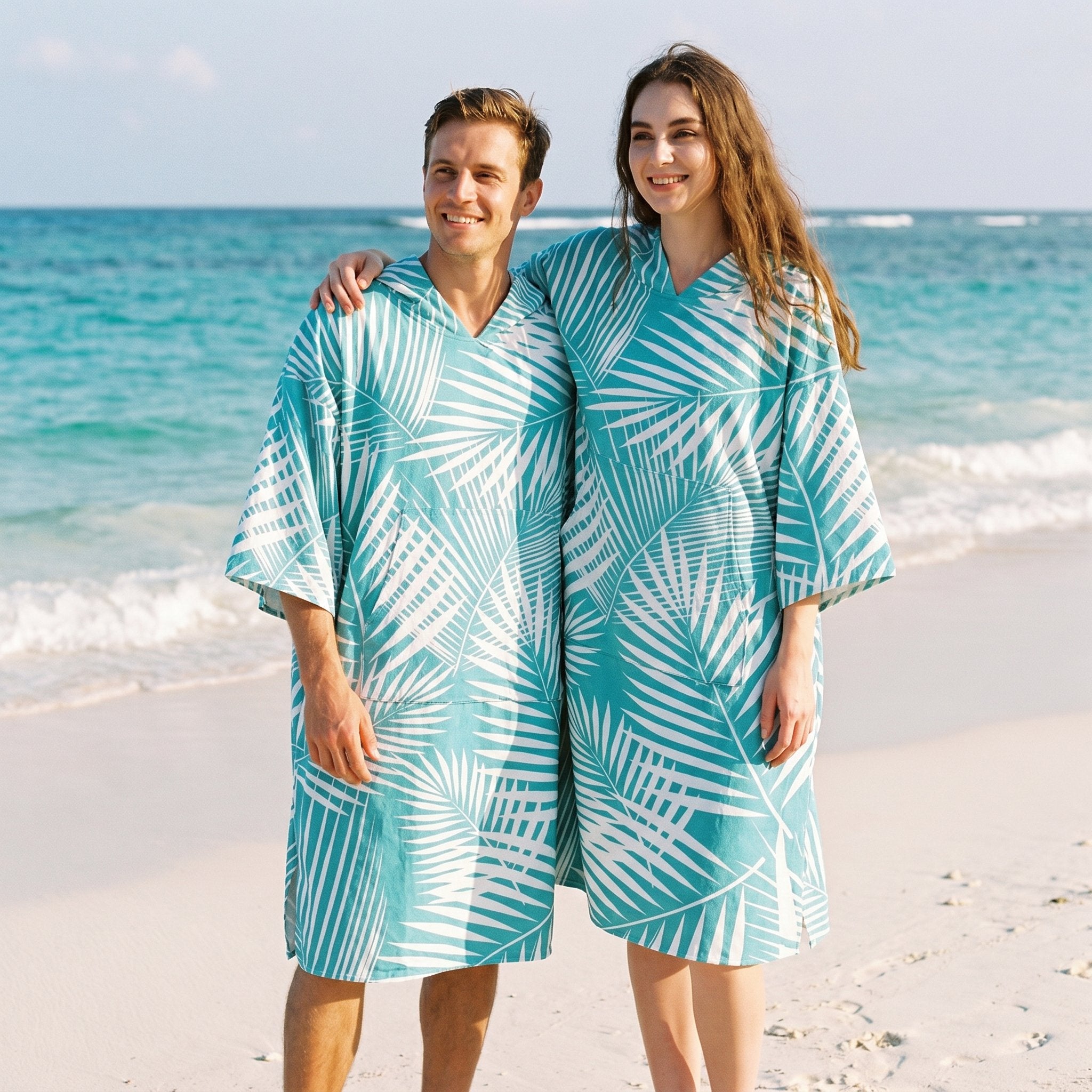 Poncho serviette à capuche motif palmes turquoise, porté par un couple souriant posant ensemble sur une plage ensoleillée.