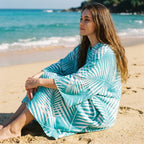 Poncho serviette à capuche motif palmes turquoise, porté par une femme assise sur le sable, le regard tourné vers l'horizon.