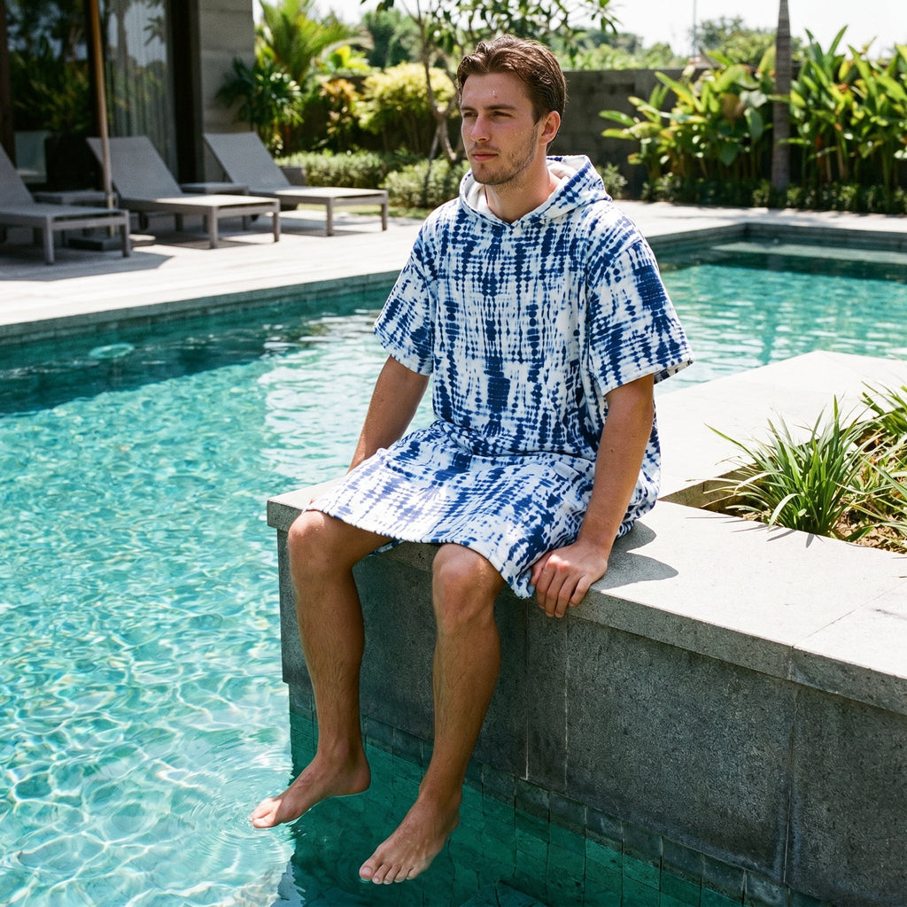 Poncho serviette a capuche motif tie-dye bleu, porté par un homme assis au bord d'une piscine, les pieds dans l'eau.