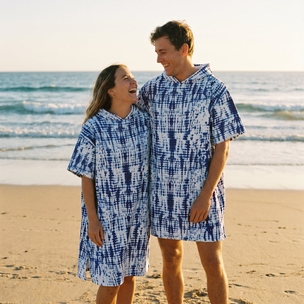 Poncho serviette a capuche motif tie-dye bleu, porté par un couple souriant sur la plage au coucher du soleil.