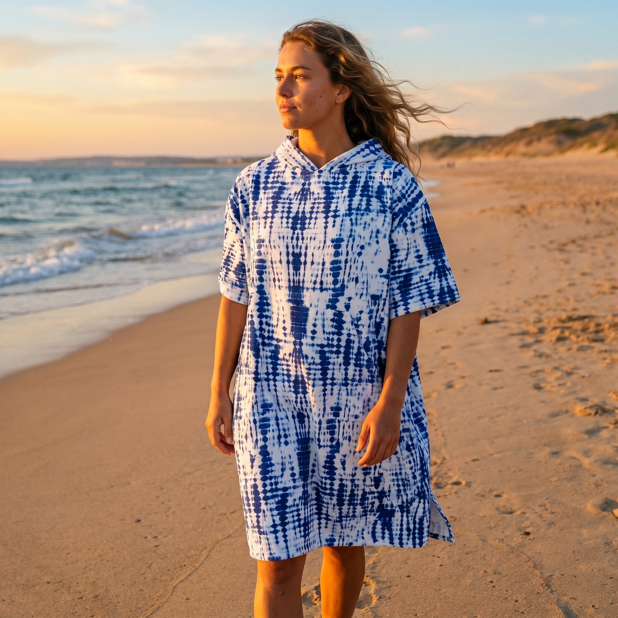 Poncho serviette a capuche motif tie-dye bleu, porté par une femme marchant seule sur le sable en bord de mer.