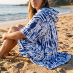 Poncho serviette a capuche motif tie-dye bleu, porté par une femme assise sur le sable, vue de dos et de côté.