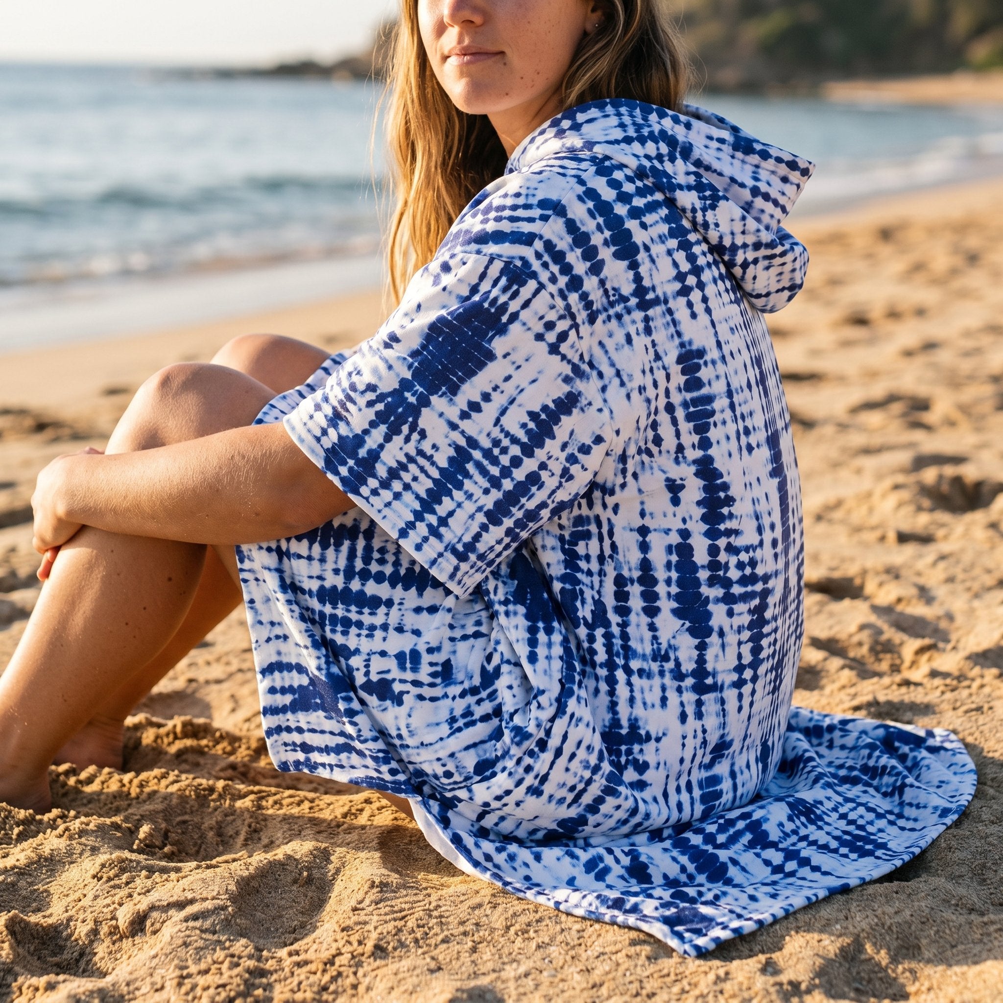 Poncho serviette a capuche motif tie-dye bleu, porté par une femme assise sur le sable, vue de dos et de côté.
