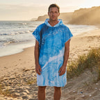 Poncho serviette a capuche motif Vague bleue, porte par un homme debout sur le sable.