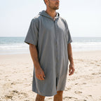 Poncho de bain zippé gris uni porté par un homme debout sur une plage de sable fin face à l'océan.