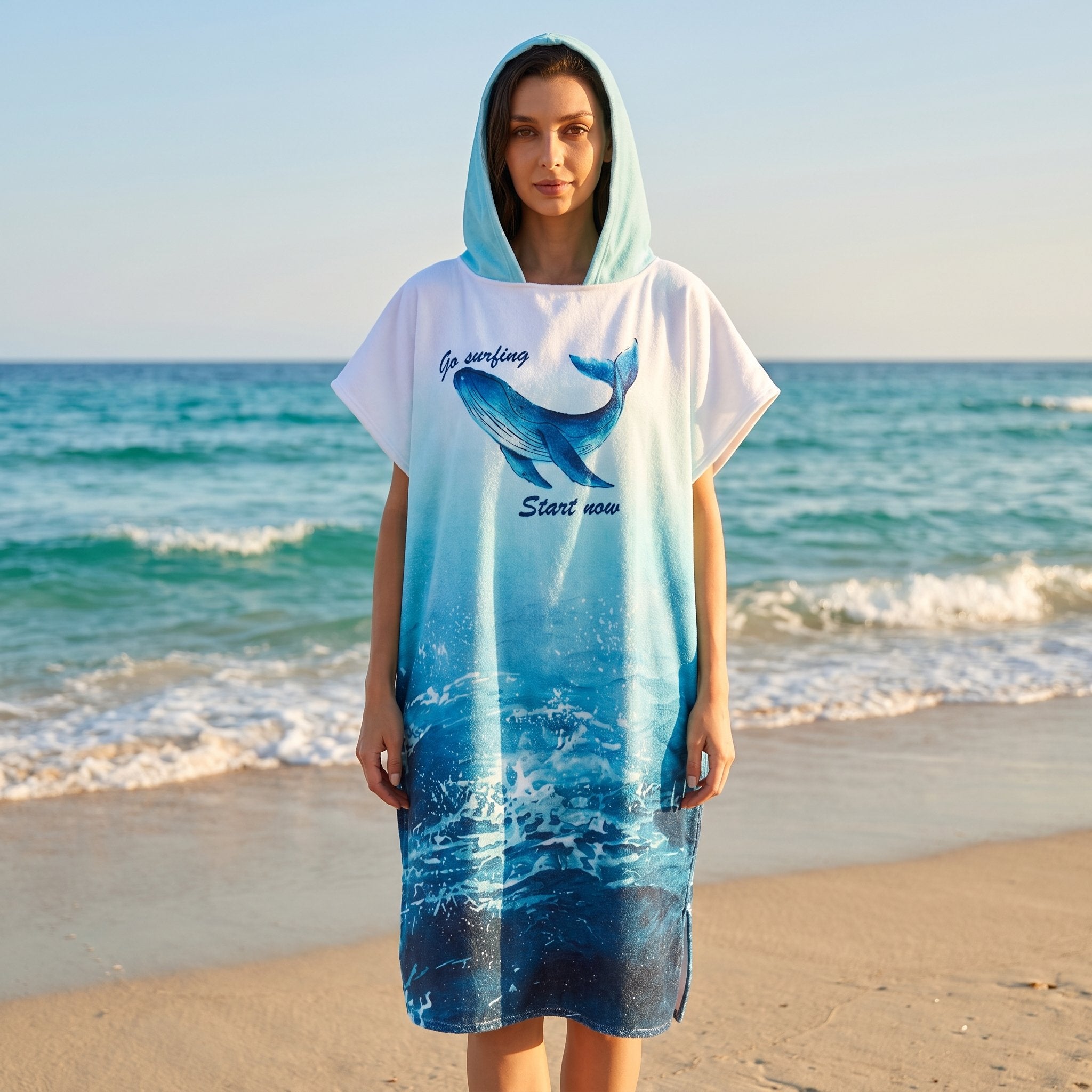 Poncho serviette à capuche motif baleine bleue, porté par une femme de face sur une plage de sable.