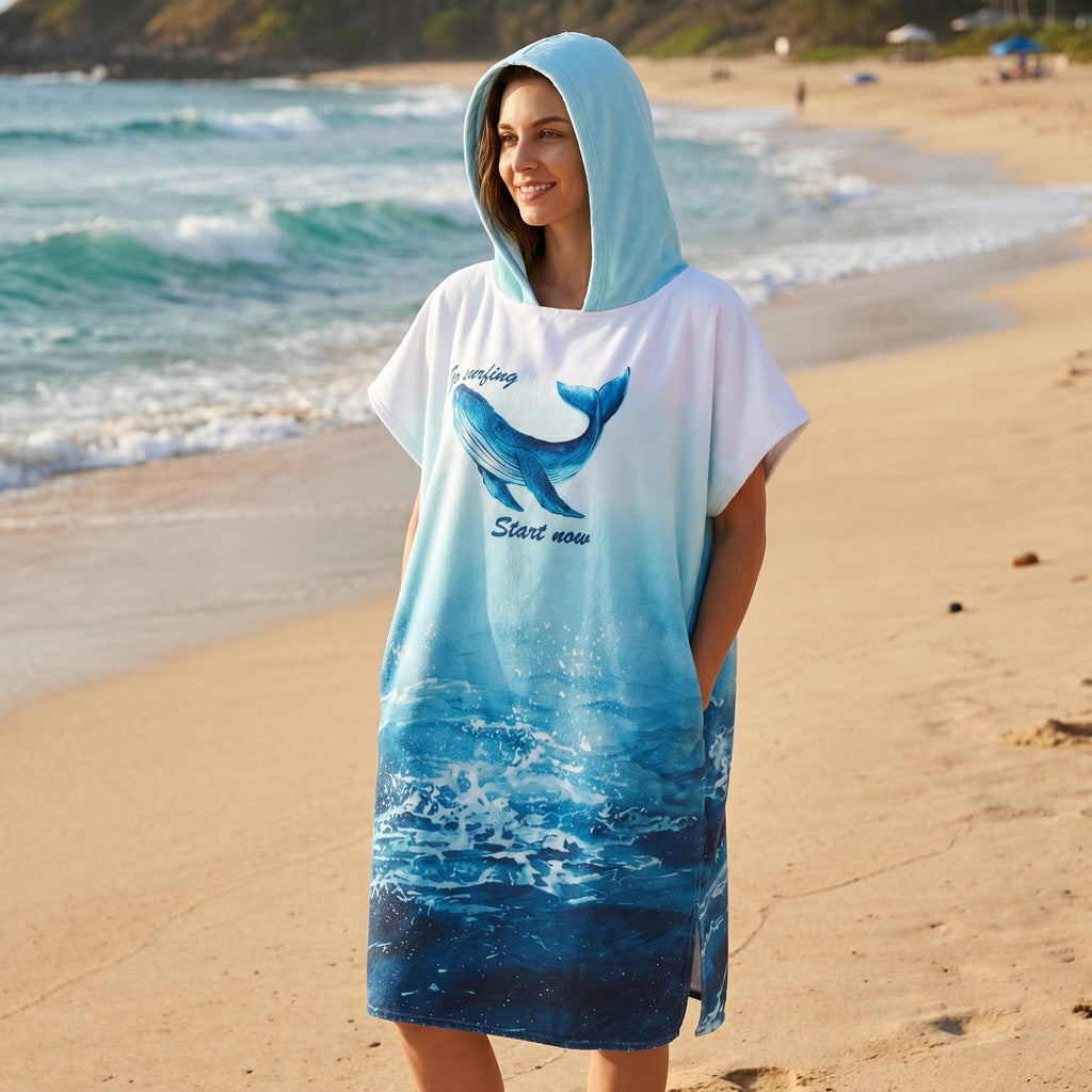 Poncho serviette à capuche motif baleine bleue, porté par une femme souriante regardant vers la droite sur la plage.
