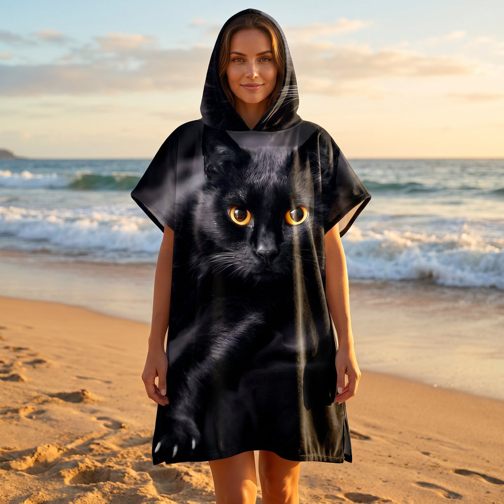 Poncho serviette à capuche motif chat noir, porté par une femme, vue de face en pied sur une plage au coucher du soleil.