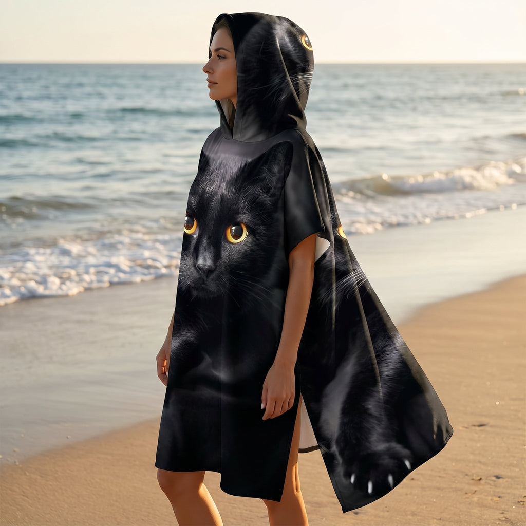 Poncho serviette à capuche motif chat noir, porté par une femme, vue de profil marchant sur le sable près de l'océan.