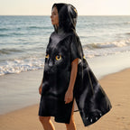 Poncho serviette à capuche motif chat noir, porté par une femme, vue de profil marchant sur le sable près de l'océan.