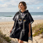 Poncho serviette à capuche motif chat noir, porté par une femme, vue de trois quarts sur une plage avec des dunes.